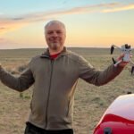 A person standing outdoors holding a drone and a remote controller, with a partially cloudy sky in the background.