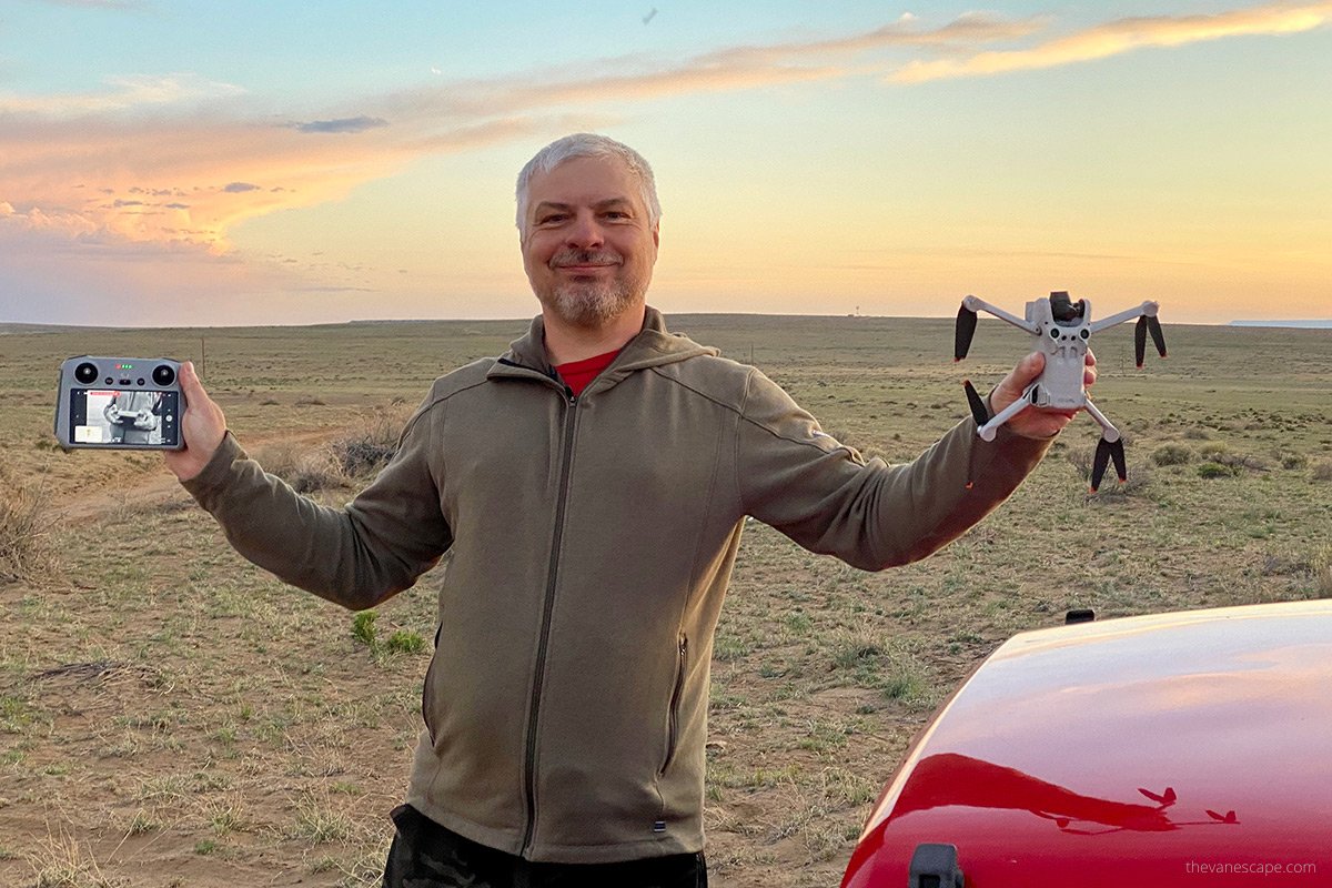 A person standing outdoors holding a drone and a remote controller, with a partially cloudy sky in the background.