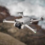 A gray quadcopter drone flies over a blurred background of rocks and water.