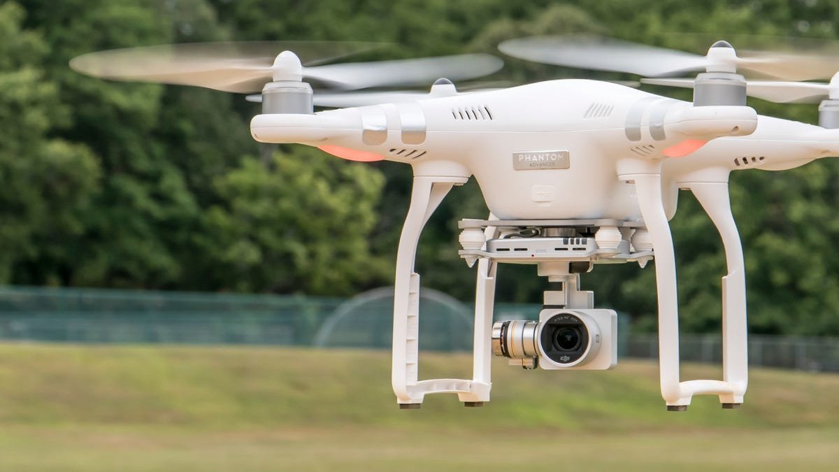 A white drone with a camera is flying outdoors, with trees and grass visible in the background.