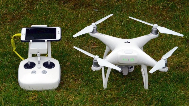 A white drone with four propellers sits on grass next to a remote control with a smartphone attached.