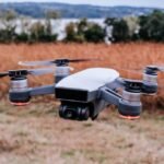 A drone hovering over a field with tall grass in the background and a body of water visible at a distance.