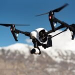 A drone with a camera hovers against a clear blue sky, with snow-capped mountains in the background.