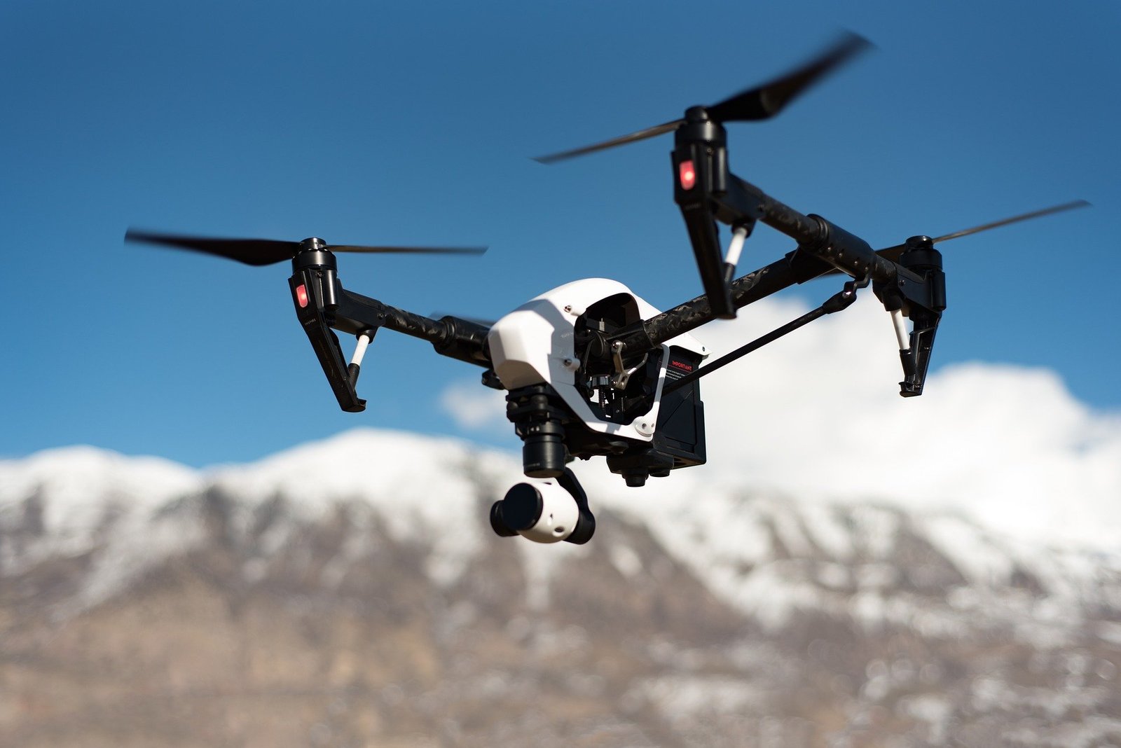 A drone with a camera hovers against a clear blue sky, with snow-capped mountains in the background.