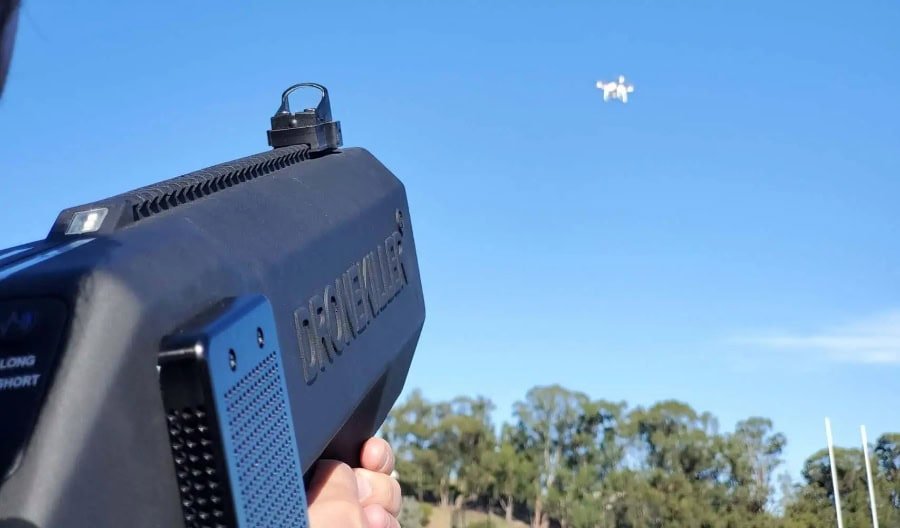 A person aims an anti-drone device at a small drone flying in the sky against a backdrop of trees.