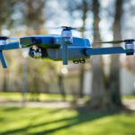 A blue drone with a camera hovers in the air against a blurred background of trees and grass.