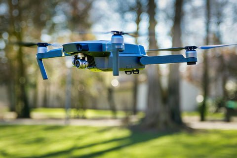 A blue drone with a camera hovers in the air against a blurred background of trees and grass.