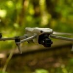 A gray drone with four propellers is flying in a green forest environment.