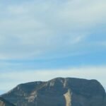 A drone flies in the sky over a rocky mountain landscape under a cloudy blue sky.