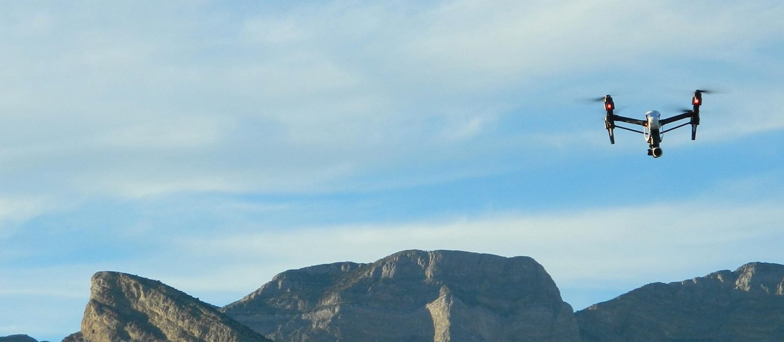 A drone flies in the sky over a rocky mountain landscape under a cloudy blue sky.