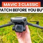 A hand holding a Mavic 3 Classic drone with a grassy field background. The text above reads "Mavic 3 Classic Watch Before You Buy!.