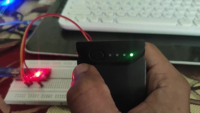 A hand holding a black device with glowing indicator lights, in front of a breadboard with a red LED light. A keyboard is visible in the background.