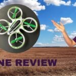 Person gesturing at a green and white drone with a rural landscape in the background. Text reads "Drone Review.