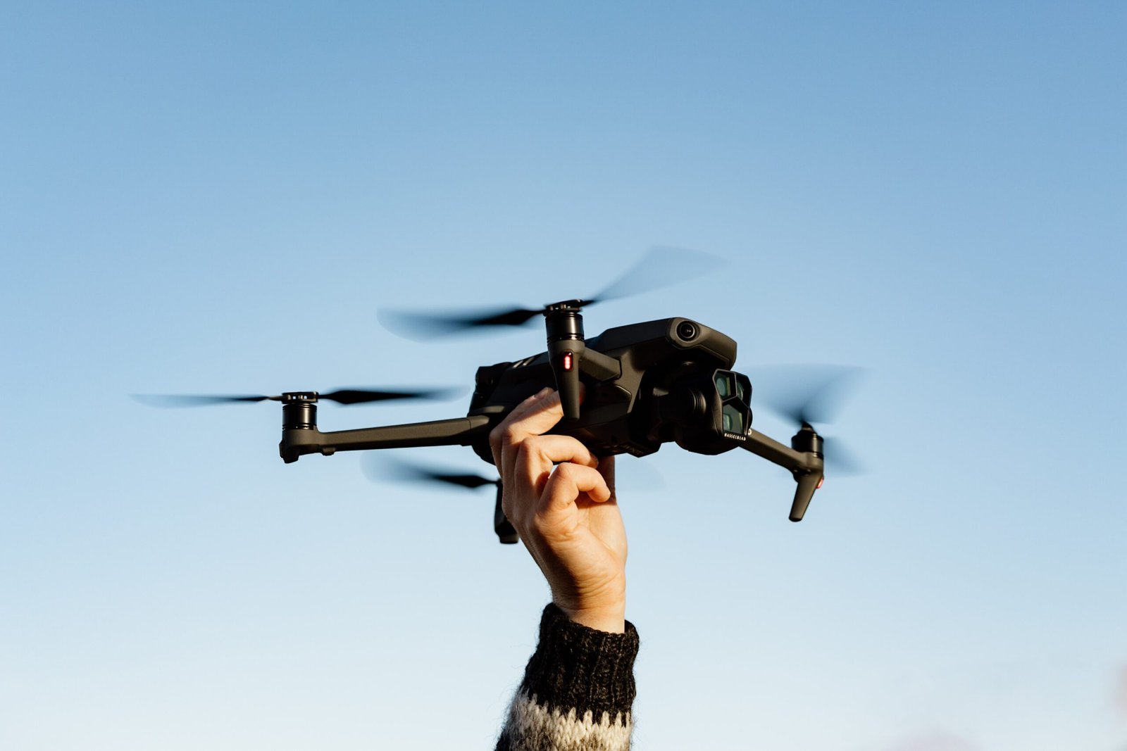 A hand holds a black drone in the air against a clear blue sky.