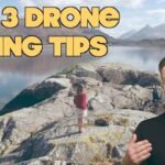 A person stands outdoors on rocks near a lake, while an inset image shows another person gesturing. Text reads "Top 3 Drone Filming Tips" and "Drone Film Guide.