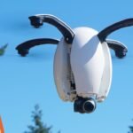 A drone is in flight against a clear blue sky. On the left, a cartoon shows aliens with a map near a smoking UFO.