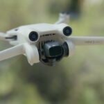 A white drone with a camera hovers mid-air outdoors, with blurred natural scenery in the background.