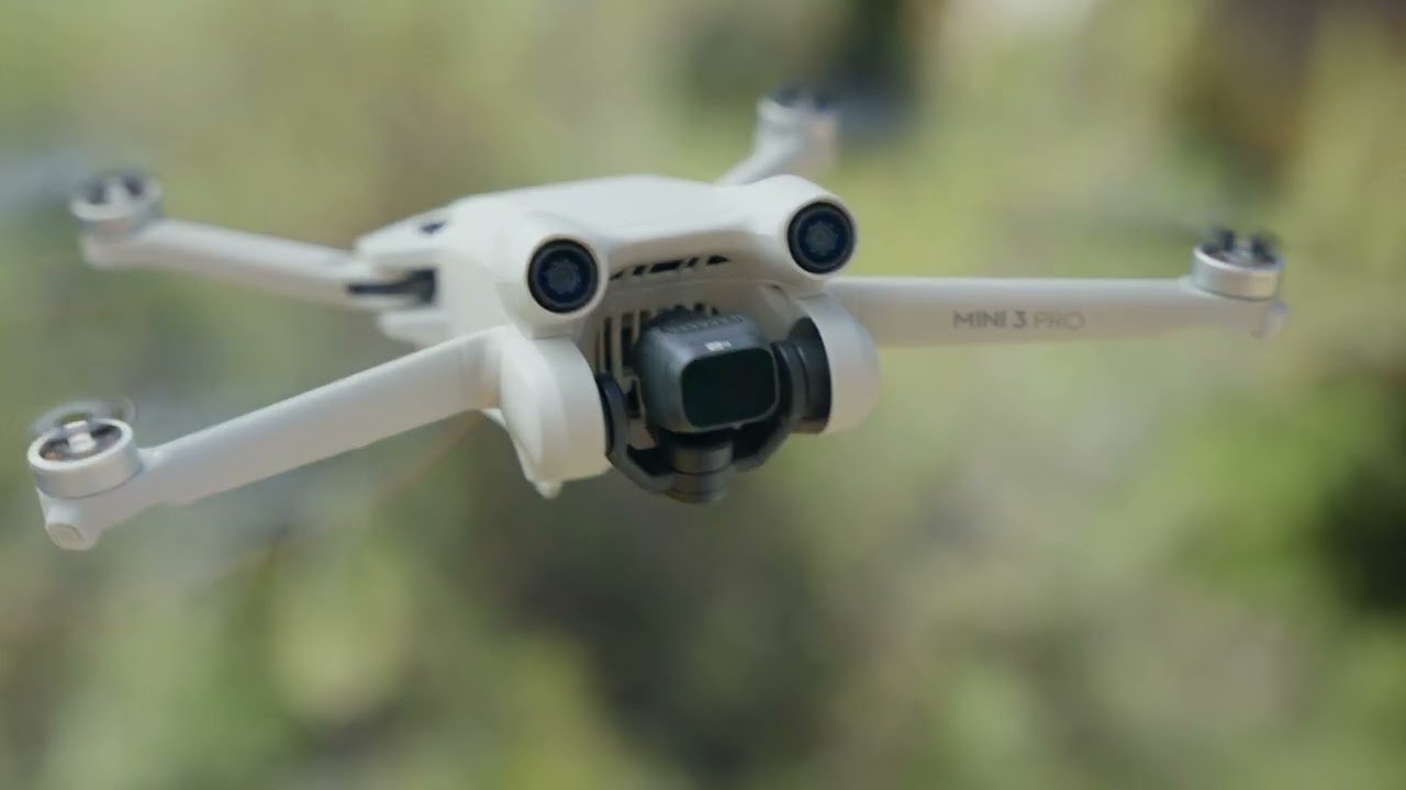 A white drone with a camera hovers mid-air outdoors, with blurred natural scenery in the background.