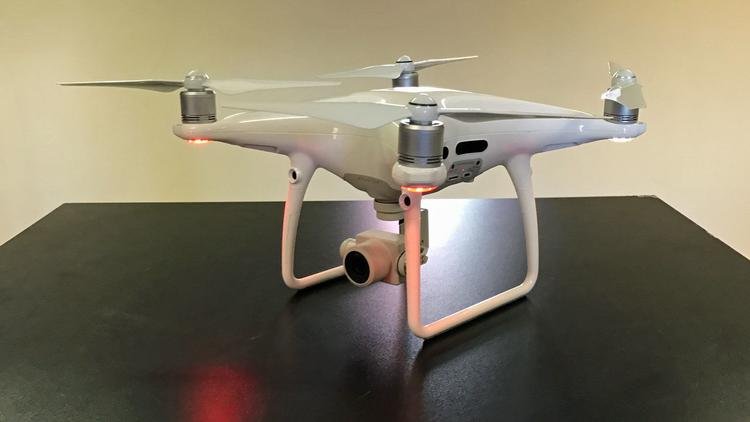 A white quadcopter drone with a mounted camera is placed on a dark surface, shown from a side angle.