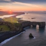 Aerial view of a coastal landscape at sunset featuring sea stacks, a large rock arch, black sand beach, and rolling green hills extending towards distant mountains.