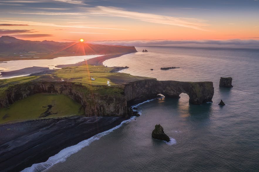 Aerial view of a coastal landscape at sunset featuring sea stacks, a large rock arch, black sand beach, and rolling green hills extending towards distant mountains.