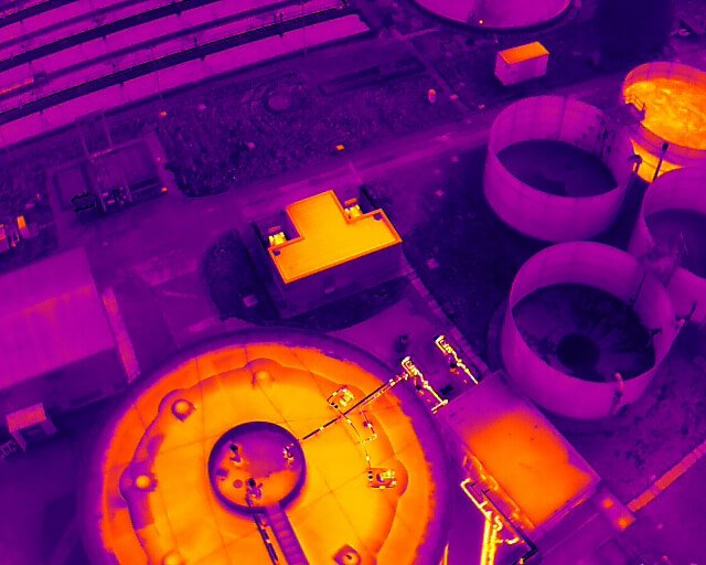 Aerial thermal image of a large industrial site with circular tanks and rectangular buildings. The colors range from purple to yellow, indicating varying temperatures.